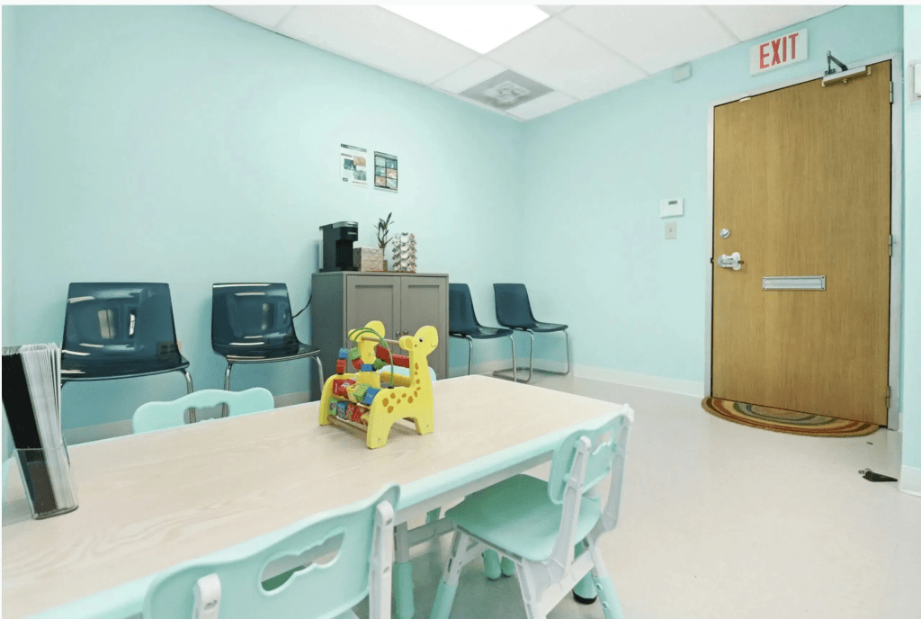 A bright and welcoming waiting room for an ABA therapy clinic, featuring a light blue wall, a table with colorful toys, and black chairs against the wall.