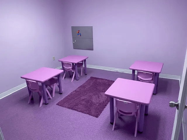 A cozy purple-themed therapy room featuring four small tables and chairs designed for children, with a soft purple rug on the floor.