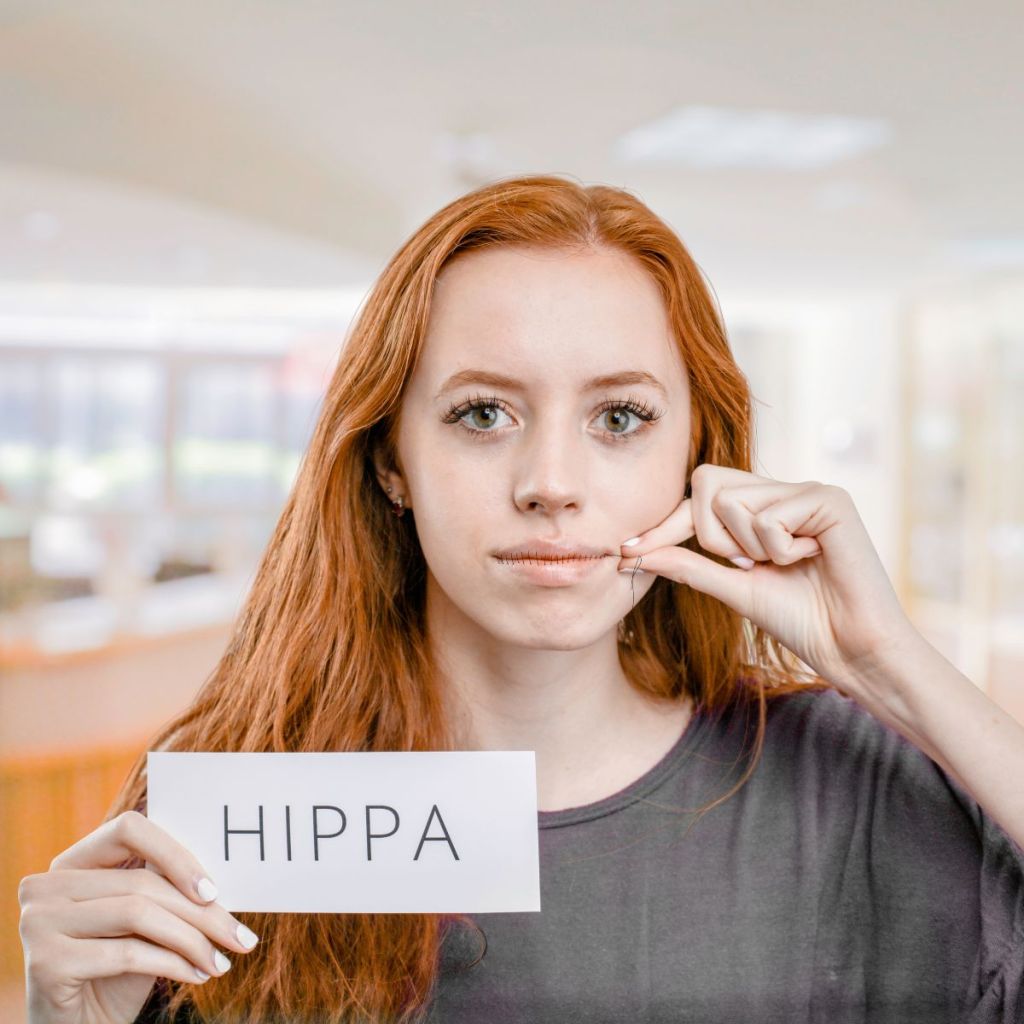 A young woman holding a sign that says 'HIPPA' while pressing her finger to her lips in a gesture of silence, set in a softly blurred background.