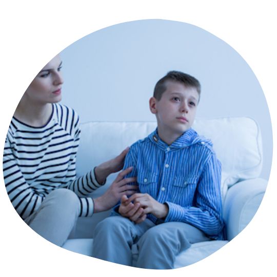 A woman is gently comforting a young boy sitting on a couch, as he looks contemplative and slightly distressed.