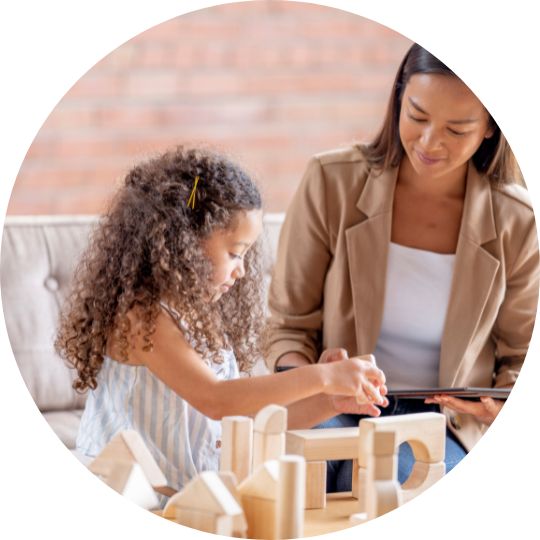A young girl with curly hair plays with wooden blocks while an adult woman, smiling and holding a tablet, observes and interacts with her.