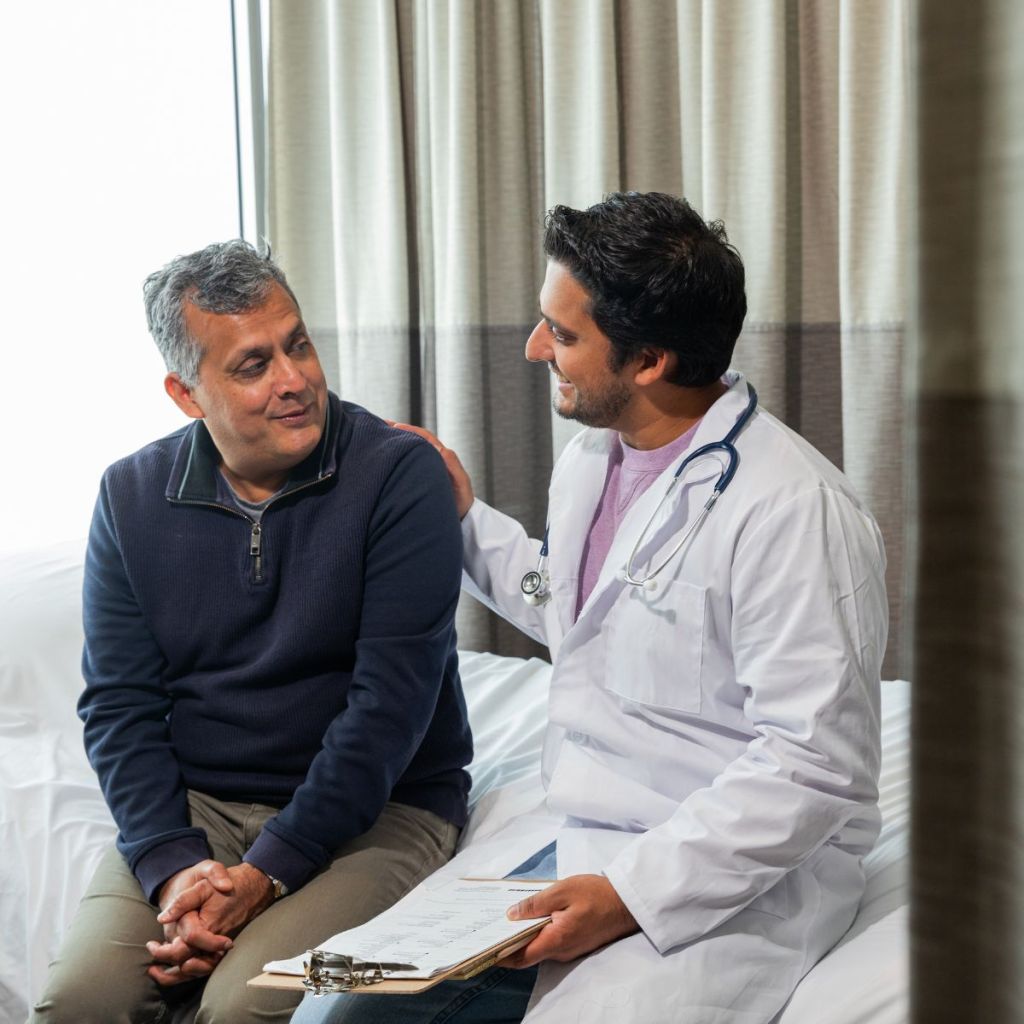 A healthcare professional in a white coat is sitting down with a patient in a medical setting, smiling and engaging in conversation. The patient appears relaxed and attentive.